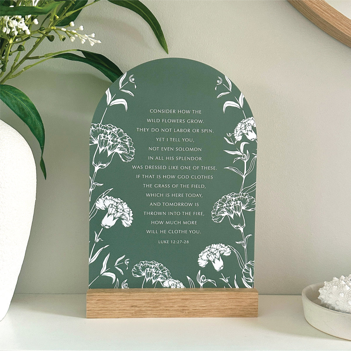 Arched Wildflowers Verse & Oak stand featuring white floral illustrations and a Bible verse from Luke 12:27-28, displayed on a handcrafted oak wooden stand on a shelf with decorative plants and a bowl in the background.